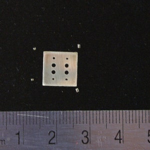 May include: A small, square, silver-coloured metal plate with six evenly spaced holes. The plate is approximately 1.3 cm wide and is surrounded by small, silver-coloured metal pieces. The plate is on a black surface.