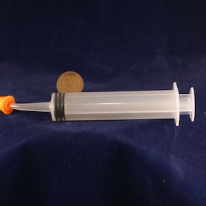 May include: A clear plastic syringe with an orange tip and black accents. The syringe is lying horizontally on a dark blue surface. A penny is next to the syringe.