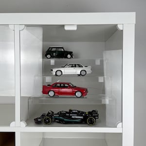 May include: A white display cabinet with four miniature toy cars. The cars are a black Mini Cooper, a white BMW, a red BMW, and a black Formula 1 race car. The cabinet has clear shelves.
