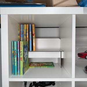 May include: A white bookshelf with multiple shelves. The top shelf has a row of colorful paperback books. The second shelf has a row of paperback books with the title "Ronald Dahl" and the third shelf has a green book with the title "Boy". The bottom shelf has a toy car and a toy truck.