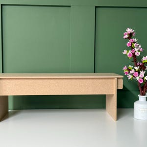 May include: A light brown, unfinished wooden bench with a rectangular top and four legs. A white vase with pink and white flowers sits to the right. The background is a green wall.