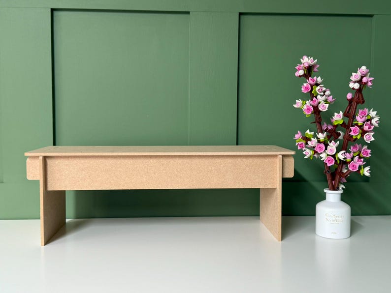 May include: A light brown, unfinished wooden bench with a rectangular top and four legs. A white vase with pink and white flowers sits to the right. The background is a green wall.