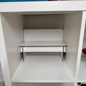 May include: A white shelf unit with a white desk in the middle section. The desk has a black top and black sides. The shelf unit has two red toy cars and a green toy car on the lower shelves.