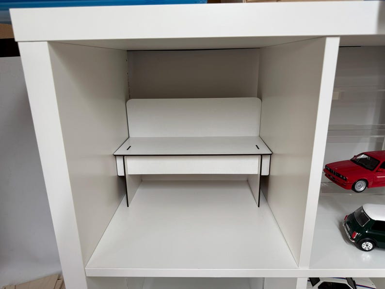 May include: A white shelf unit with a white desk in the middle section. The desk has a black top and black sides. The shelf unit has two red toy cars and a green toy car on the lower shelves.