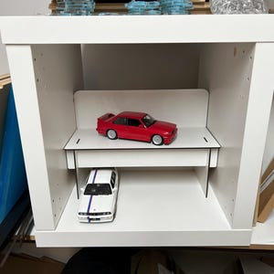 May include: A white shelf unit with two levels, each holding a red and white toy car. The shelf unit is made of wood and has a simple design.