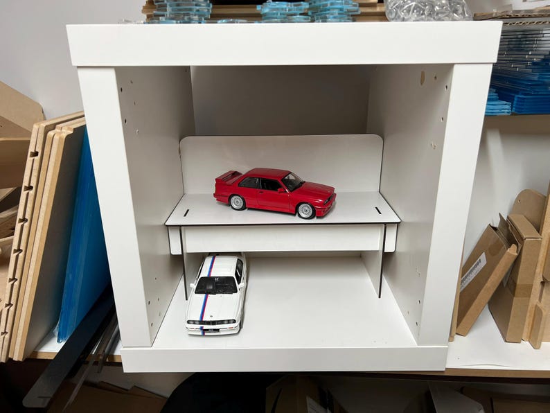 May include: A white shelf unit with two levels, each holding a red and white toy car. The shelf unit is made of wood and has a simple design.