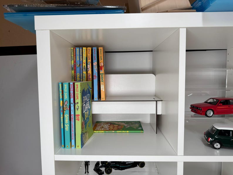 May include: White bookshelf with multiple shelves. The top shelf has a row of colorful paperback books with the title "Ronald Dahl" on several of the books. The second shelf has a row of colorful paperback books with the title "Going Solo" on one of the books. The third shelf has a green and yellow paperback book. The bottom shelf has a black toy car. The right side of the bookshelf has a red toy car and a green toy car.