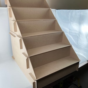May include: A multi-tiered, unfinished wooden display shelf. The shelf has five stepped levels, each with a small lip to hold items. The construction appears to be made of medium-density fibreboard (MDF). The display is designed to be freestanding.