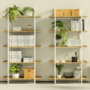 Puede incluir: Dos estanter&iacute;as de metal blanco con estantes de madera, llenas de libros, plantas y objetos decorativos. Las estanter&iacute;as est&aacute;n contra una pared blanca con una l&aacute;mpara de pie de tr&iacute;pode negra a la izquierda.
