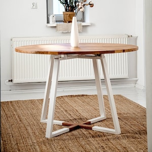 May include: A round wooden table with white legs and a white vase with cotton flowers on top. The table is on a brown rug in a white room.