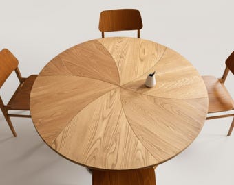 May include: A round wooden dining table with a light brown finish, surrounded by four matching chairs. The table's surface is divided into pie-shaped sections. A small white vase with a pen is on the table.