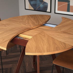 Op de afbeelding: Een uitschuifbare eettafel in lichtbruin, met een opvallende halfronde vorm. Het houten oppervlak vertoont een duidelijk nerfpatroon, ondersteund door een donkerrood metalen onderstel. Twee bruine stoelen omringen de tafel.