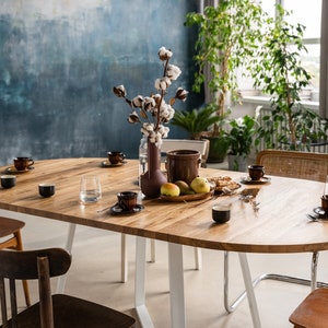 K&ouml;nnte beinhalten: Ein ovaler Esstisch aus Holz mit wei&szlig;en Metallbeinen, gedeckt f&uuml;r eine Mahlzeit mit braunen Keramikbechern, Tellern und Besteck. Eine Vase mit einem Baumwollzweig steht in der Mitte des Tisches, zusammen mit einer Schale Obst und einem Krug.