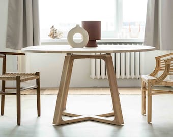 Könnte beinhalten: Ein runder Esstisch aus hellem Holz mit einem einzigartigen Gestell, flankiert von zwei Stühlen. Ein Stuhl hat einen gewebten Sitz und einen dunklen Holzrahmen, der andere ist aus hellem Webmaterial gefertigt. Dekorative Vasen stehen auf dem Tisch.