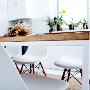 May include: A modern dining table with a light wood top and white metal legs. White chairs with wooden legs are positioned around the table. A dried floral arrangement sits on the table. The scene is in a bright, white kitchen.
