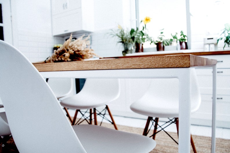 May include: A modern dining table with a light wood top and white metal legs. White chairs with wooden legs are positioned around the table. A dried floral arrangement sits on the table. The scene is in a bright, white kitchen.