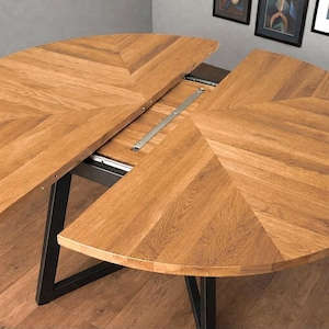 Op de afbeelding: Een ronde, uitschuifbare houten tafel met een lichtbruine afwerking. Het tafelblad is verdeeld in twee halve cirkelvormige secties met een visgraatpatroon. De tafel heeft een zwart metalen onderstel en is gedeeltelijk uitgeschoven.