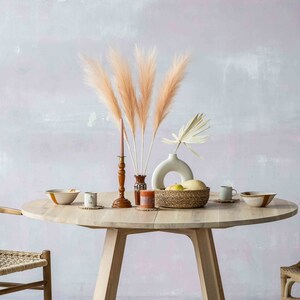 May include: A round wooden table with two chairs, set with a bowl, a vase with dried pampas grass, a candle, and a basket of fruit. The table is in a room with a light pink wall and a macrame wall hanging.