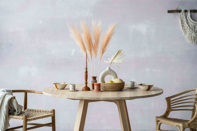 May include: A round wooden table with two chairs, set with a bowl, a vase with dried pampas grass, a candle, and a basket of fruit. The table is in a room with a light pink wall and a macrame wall hanging.