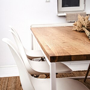 May include: A modern dining table with a light wood grain top and white metal legs. Three white plastic chairs are positioned around the table. A bundle of dried wheat stalks rests on the table. The room has white walls and a neutral rug.