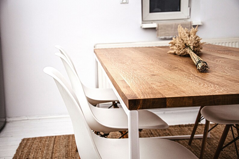 May include: A modern dining table with a light wood grain top and white metal legs. Three white plastic chairs are positioned around the table. A bundle of dried wheat stalks rests on the table. The room has white walls and a neutral rug.