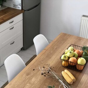 May include: A wooden table with a wire basket filled with apples, oranges, and avocados. There are two corn cobs on the table and a white chair in the background.