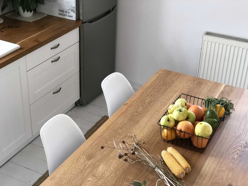 May include: A wooden table with a wire basket filled with apples, oranges, and avocados. There are two corn cobs on the table and a white chair in the background.