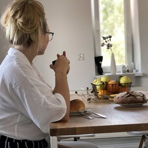 May include: A woman sits at a table with a cup in her hand, looking out a window. The table is set with a loaf of bread, a bowl of fruit, and a few sprigs of dried flowers.