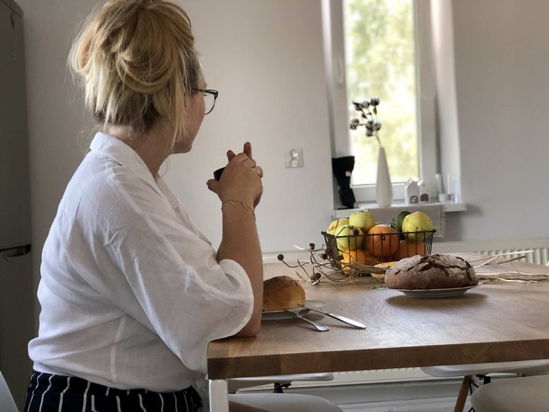 May include: A woman sits at a table with a cup in her hand, looking out a window. The table is set with a loaf of bread, a bowl of fruit, and a few sprigs of dried flowers.