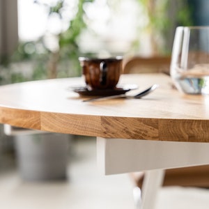 K&ouml;nnte beinhalten: Nahaufnahme einer runden Tischplatte aus hellem Holz mit einem wei&szlig;en Untergestell. Die Tischplatte hat eine nat&uuml;rliche Holzmaserung. Ein Glas Wasser und eine Kaffeetasse sind im Hintergrund unscharf.