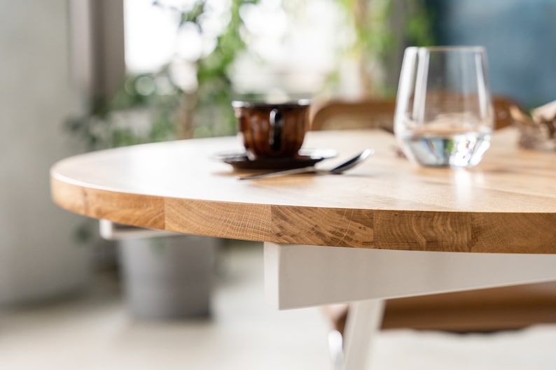 K&ouml;nnte beinhalten: Nahaufnahme einer runden Tischplatte aus hellem Holz mit einem wei&szlig;en Untergestell. Die Tischplatte hat eine nat&uuml;rliche Holzmaserung. Ein Glas Wasser und eine Kaffeetasse sind im Hintergrund unscharf.