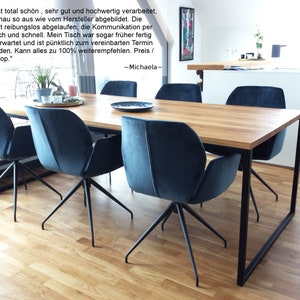 May include: A wooden dining table with a black metal frame and four blue velvet chairs. The table is set with a black plate and a glass on each chair. The table is in a modern dining room with a white kitchen in the background.