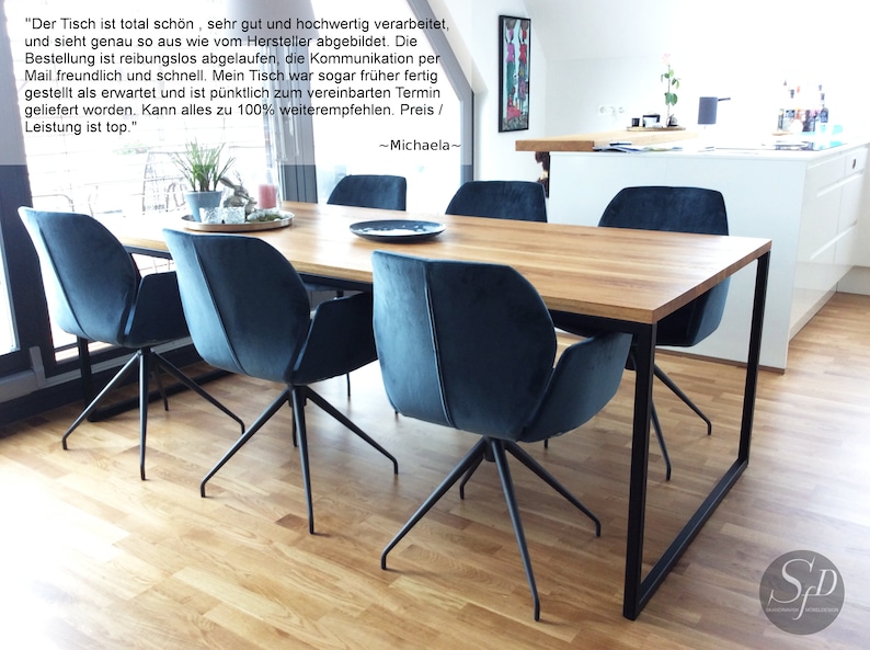May include: A wooden dining table with a black metal frame and four blue velvet chairs. The table is set with a black plate and a glass on each chair. The table is in a modern dining room with a white kitchen in the background.