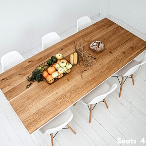 May include: A rectangular wooden dining table with a light brown finish, surrounded by six white chairs. A black wire basket holds fruit, and a loaf of bread sits on the table. The table is approximately 72 inches long.