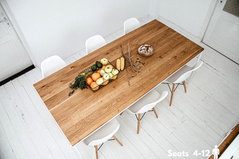 May include: A rectangular wooden dining table with a light brown finish, surrounded by six white chairs. A black wire basket holds fruit, and a loaf of bread sits on the table. The table is approximately 72 inches long.