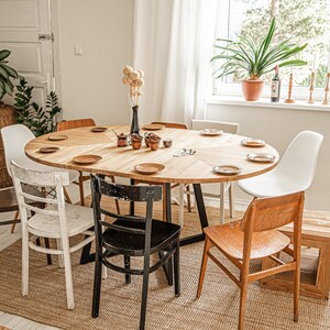 May include: A wooden oval dining table with six chairs, set for a meal. The table has a light wood finish and is surrounded by chairs in various styles, including white, black, and natural wood. The table is set with plates, cutlery, and a vase of dried flowers.