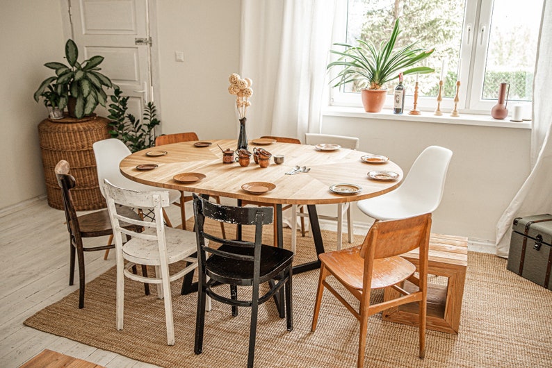 May include: A wooden oval dining table with six chairs, set for a meal. The table has a light wood finish and is surrounded by chairs in various styles, including white, black, and natural wood. The table is set with plates, cutlery, and a vase of dried flowers.