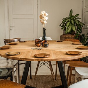 May include: A round wooden dining table with black metal legs, surrounded by chairs in various styles and colors. The table is set with brown plates and a vase with dried flowers.