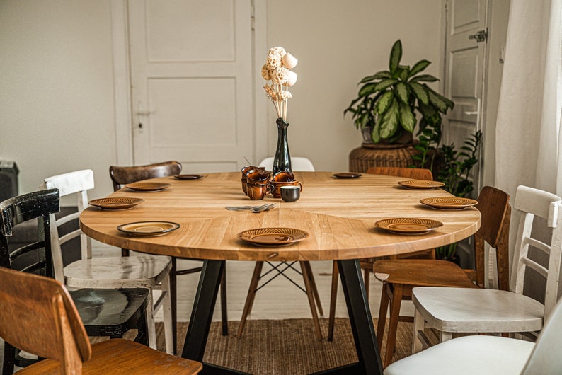 May include: A round wooden dining table with black metal legs, surrounded by chairs in various styles and colors. The table is set with brown plates and a vase with dried flowers.