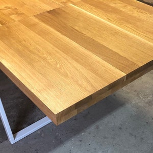 May include: A close-up of a wooden table top with a white metal frame. The table top is made of light wood planks and has a natural finish. The table is in a room with a concrete floor.