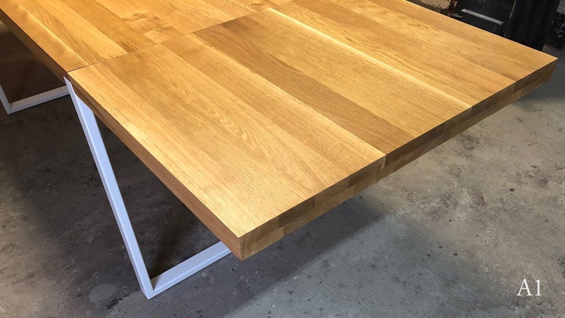 May include: A close-up of a wooden table top with a white metal frame. The table top is made of light wood planks and has a natural finish. The table is in a room with a concrete floor.