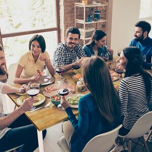 May include: A group of people seated around a wooden table, enjoying a meal. The table is set with plates, glasses of red wine, and various dishes. The scene suggests a social gathering or a dinner party.