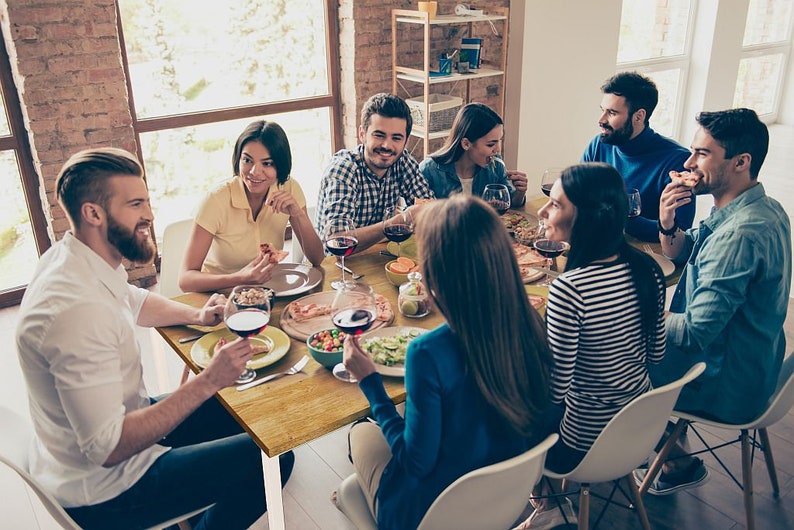 May include: A group of people seated around a wooden table, enjoying a meal. The table is set with plates, glasses of red wine, and various dishes. The scene suggests a social gathering or a dinner party.