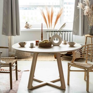May include: A round wooden dining table with a natural wood finish and a cross-shaped base. Two chairs are placed around the table, one with a woven seat and the other with a woven back and seat. A vase with dried pampas grass sits on the table, along with a bowl of fruit and candles.