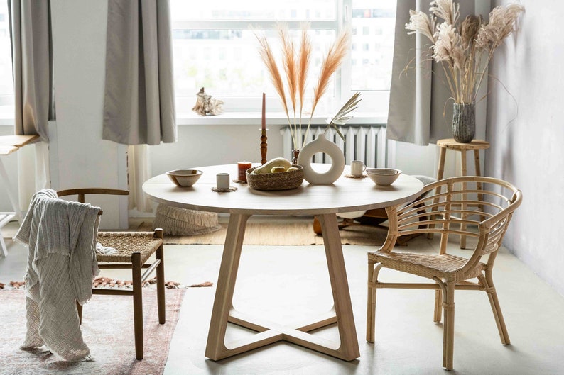 May include: A round wooden dining table with a natural wood finish and a cross-shaped base. Two chairs are placed around the table, one with a woven seat and the other with a woven back and seat. A vase with dried pampas grass sits on the table, along with a bowl of fruit and candles.