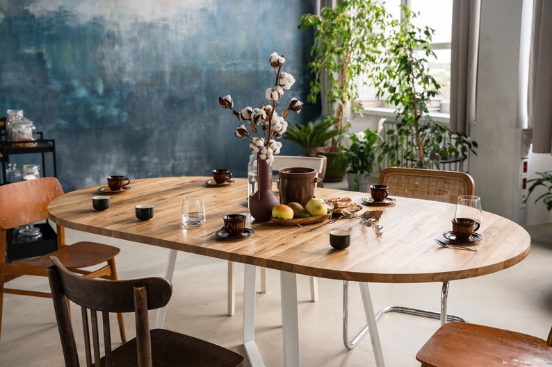 K&ouml;nnte beinhalten: Ein ovaler Esstisch aus Holz mit wei&szlig;en Metallbeinen, gedeckt f&uuml;r eine Mahlzeit mit braunen Keramikbechern, Tellern und Besteck. Eine Vase mit einem Baumwollzweig steht in der Mitte des Tisches, zusammen mit einer Schale Obst und einem Krug.