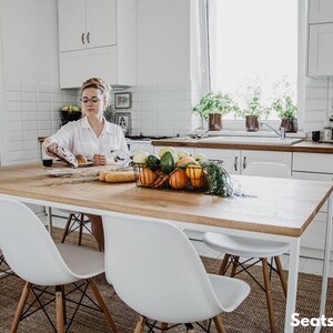 May include: A bright kitchen scene with a wooden dining table and four white chairs. A woman is preparing food. A wire basket on the table holds vegetables. The room has white cabinets and a window with plants. The table seats 4-12 people.