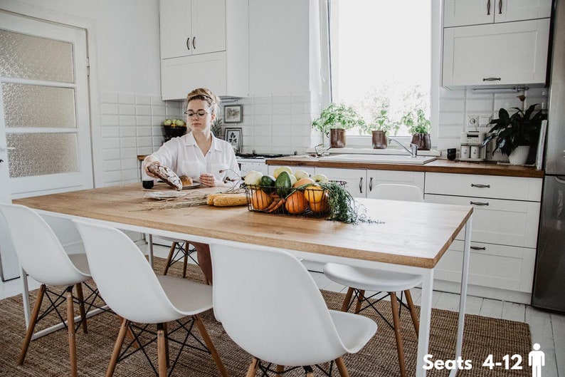 May include: A bright kitchen scene with a wooden dining table and four white chairs. A woman is preparing food. A wire basket on the table holds vegetables. The room has white cabinets and a window with plants. The table seats 4-12 people.