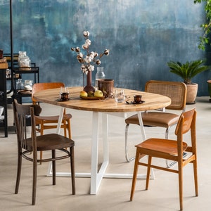 K&ouml;nnte beinhalten: Ein ovaler Esstisch aus Holz mit einem wei&szlig;en Metallgestell, umgeben von vier St&uuml;hlen. Der Tisch ist mit einer Vase mit Baumwollblumen, Obst und Gl&auml;sern gedeckt. Die St&uuml;hle sind in verschiedenen Stilen gehalten, darunter ein brauner Holzstuhl, ein brauner Stuhl mit einem geflochtenen Sitz und ein Stuhl mit einem verchromten Gestell.
