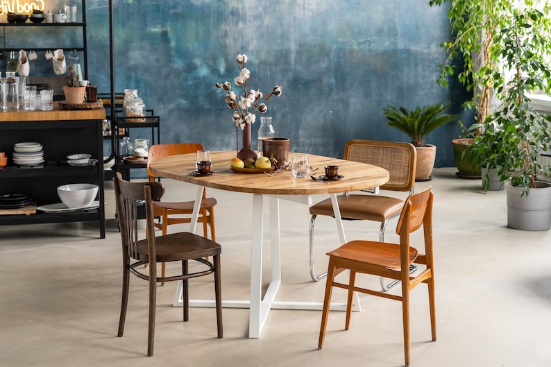 K&ouml;nnte beinhalten: Ein ovaler Esstisch aus Holz mit einem wei&szlig;en Metallgestell, umgeben von vier St&uuml;hlen. Der Tisch ist mit einer Vase mit Baumwollblumen, Obst und Gl&auml;sern gedeckt. Die St&uuml;hle sind in verschiedenen Stilen gehalten, darunter ein brauner Holzstuhl, ein brauner Stuhl mit einem geflochtenen Sitz und ein Stuhl mit einem verchromten Gestell.